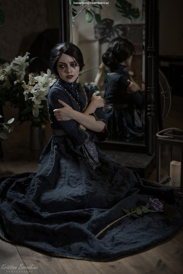 A woman dressed in a dark, vintage-style gown sits on the floor, hugging her knees with a contemplative expression. She is surrounded by indoor plants and a mirror reflecting her image. A purple rose lies beside her on the ground.