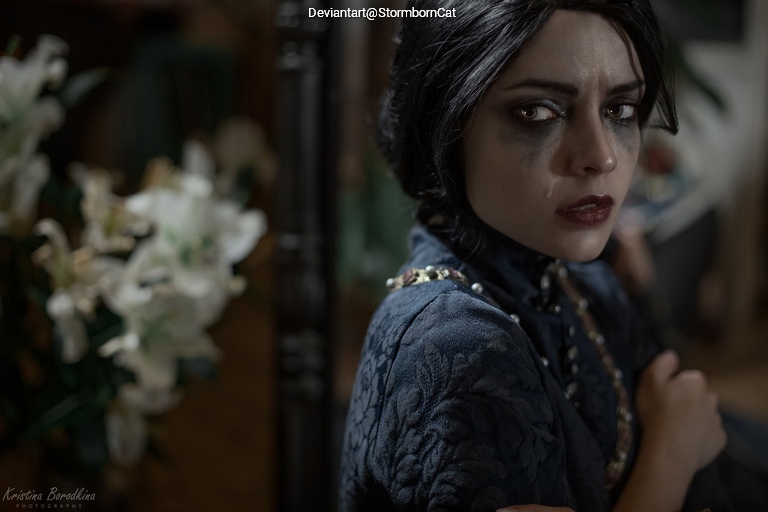 A close-up portrait of a woman with dark hair and dramatic makeup, wearing a patterned blue dress, looking over her shoulder with a serious expression, surrounded by white flowers in a softly lit setting.