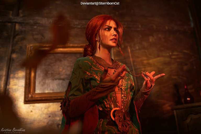 A woman with red hair wearing a green and ornate cloak, standing dramatically in a dimly lit room with golden accents.