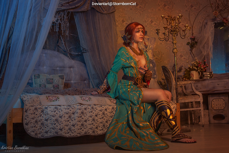A woman in a vibrant, intricate outfit sits on a bed in a cozy, softly lit room, decorated with floral patterns and vintage furniture.