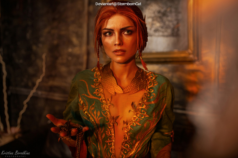 A woman with red hair is wearing a green outfit with intricate designs and a gold necklace, looking thoughtfully at the camera in a dimly lit room.