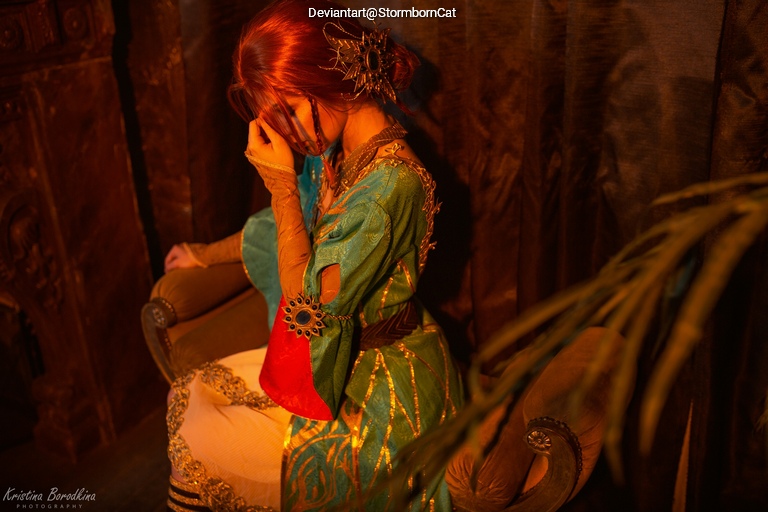 A woman with long red hair wearing a colorful historical costume is seated on a chair, covering her face with one hand in a dramatic pose, surrounded by dark fabric and shadows.