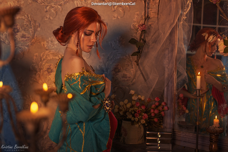 A woman with long red hair in an elegant turquoise dress, standing in a softly lit room adorned with floral wallpaper and candles. A reflection of her can be seen in a nearby mirror.