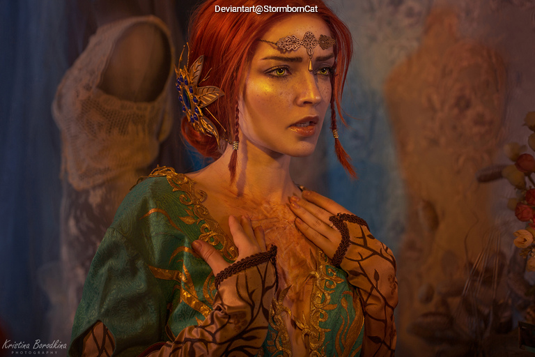 A woman with red hair wearing a blue and gold costume, adorned with a decorative hairpiece and intricate makeup, showing an emotional expression against a soft, artistic backdrop.