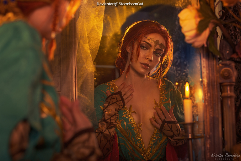 A woman with red hair styled in elaborate braids gazes at her reflection in a mirror, dressed in a colorful, intricate costume adorned with patterns. Soft lighting and a candle flame illuminate the scene, highlighting her expressive features and detailed makeup.
