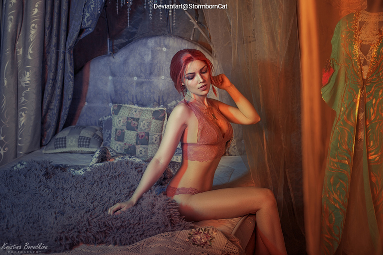 A woman with red hair sitting on a bed, wearing a lace lingerie set, surrounded by decorative pillows and soft fabrics. The lighting creates a warm and intimate atmosphere.