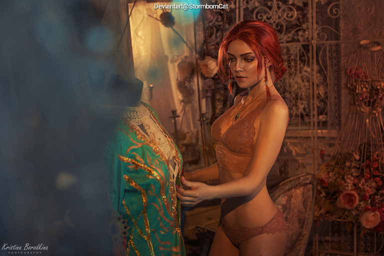 A woman with red hair in lingerie examines a colorful, ornate dress on a mannequin in a softly lit, vintage-themed room.