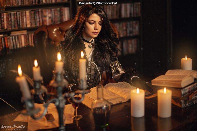A woman with long dark hair sits at a wooden table surrounded by candles and books, deeply focused on reading an open book.