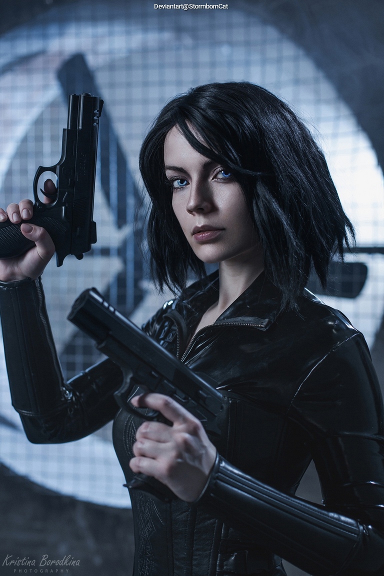 A woman with short black hair poses confidently holding two handguns, dressed in a sleek black outfit, against a textured backdrop.
