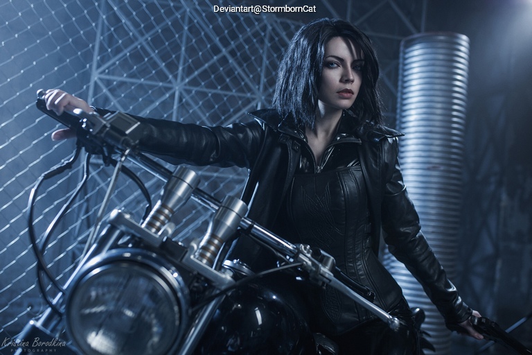 A person in a black leather outfit poses confidently on a motorcycle, set against an industrial backdrop with a chain-link fence.