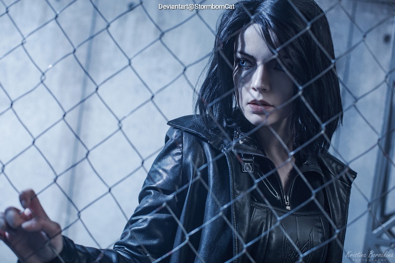A person with dark hair dressed in a black leather jacket is standing by a chain-link fence, looking intently.