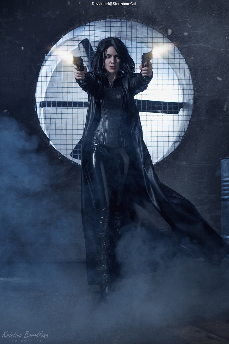 A woman in a black leather outfit stands confidently, holding two guns and aiming them forward. She is surrounded by mist and stands in front of a large circular window with a grid pattern.