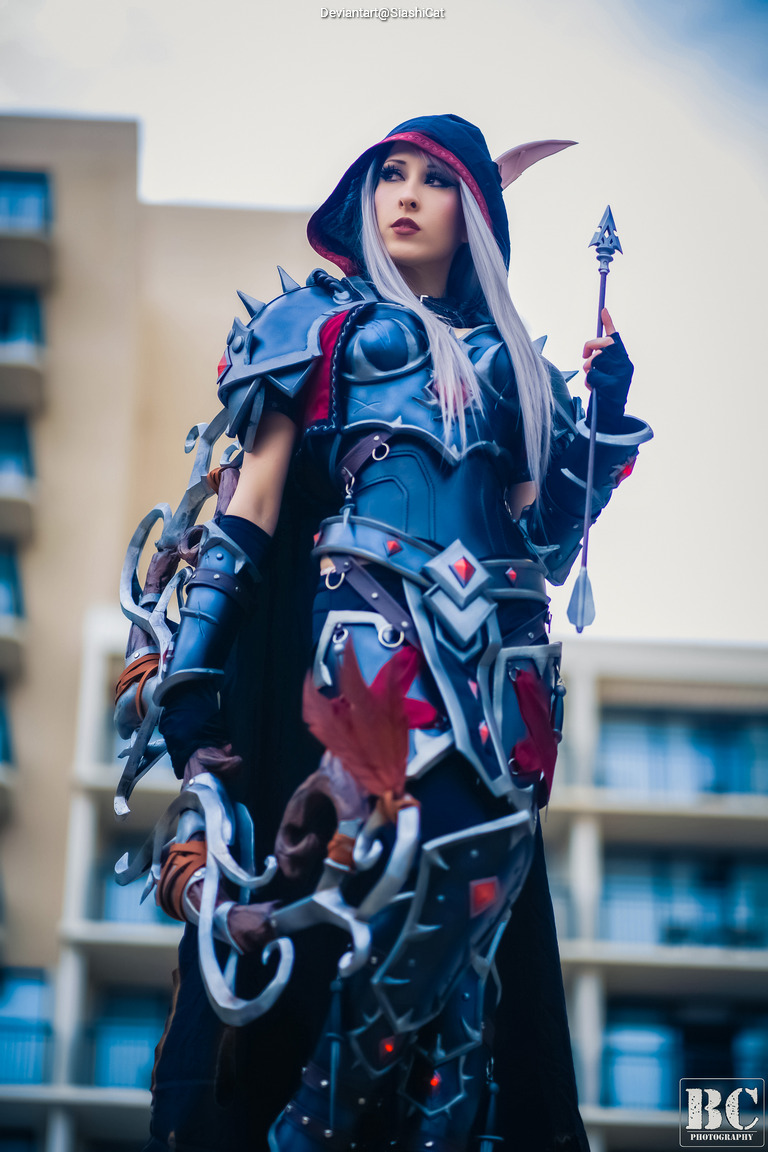 Cosplayer dressed in intricate dark armor with a hood, posing confidently and holding a spear, against a blurred building backdrop.