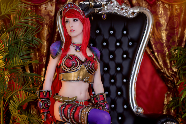 A woman in a colorful fantasy costume with red hair and horned headpiece, sitting on an ornate black throne against a luxurious backdrop with greenery.