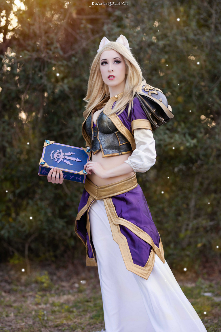 A woman in a fantasy costume holds a book, featuring purple and gold attire with a fitted top and flowing white skirt, standing outdoors with greenery in the background.