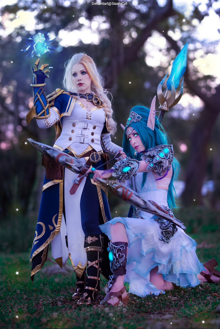 Two female cosplayers pose in a forest setting, one in a detailed white and gold costume holding a glowing blue orb, and the other in a flowing white dress with turquoise hair, kneeling and grasping a weapon with glowing accents.