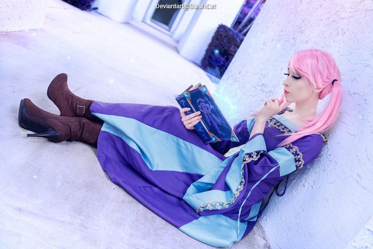 A person with pink hair wearing a purple and blue dress is sitting on the ground, holding a book while looking contemplatively to the side.