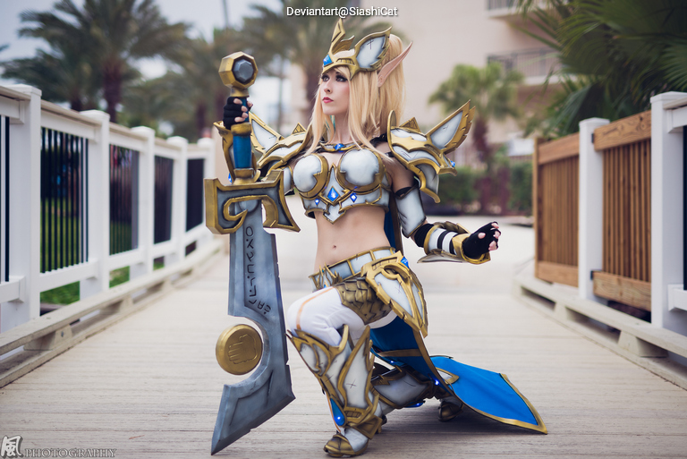 A female cosplayer dressed in elaborate armored costume, resembling a fantasy character, kneeling on a wooden pathway with palm trees in the background.