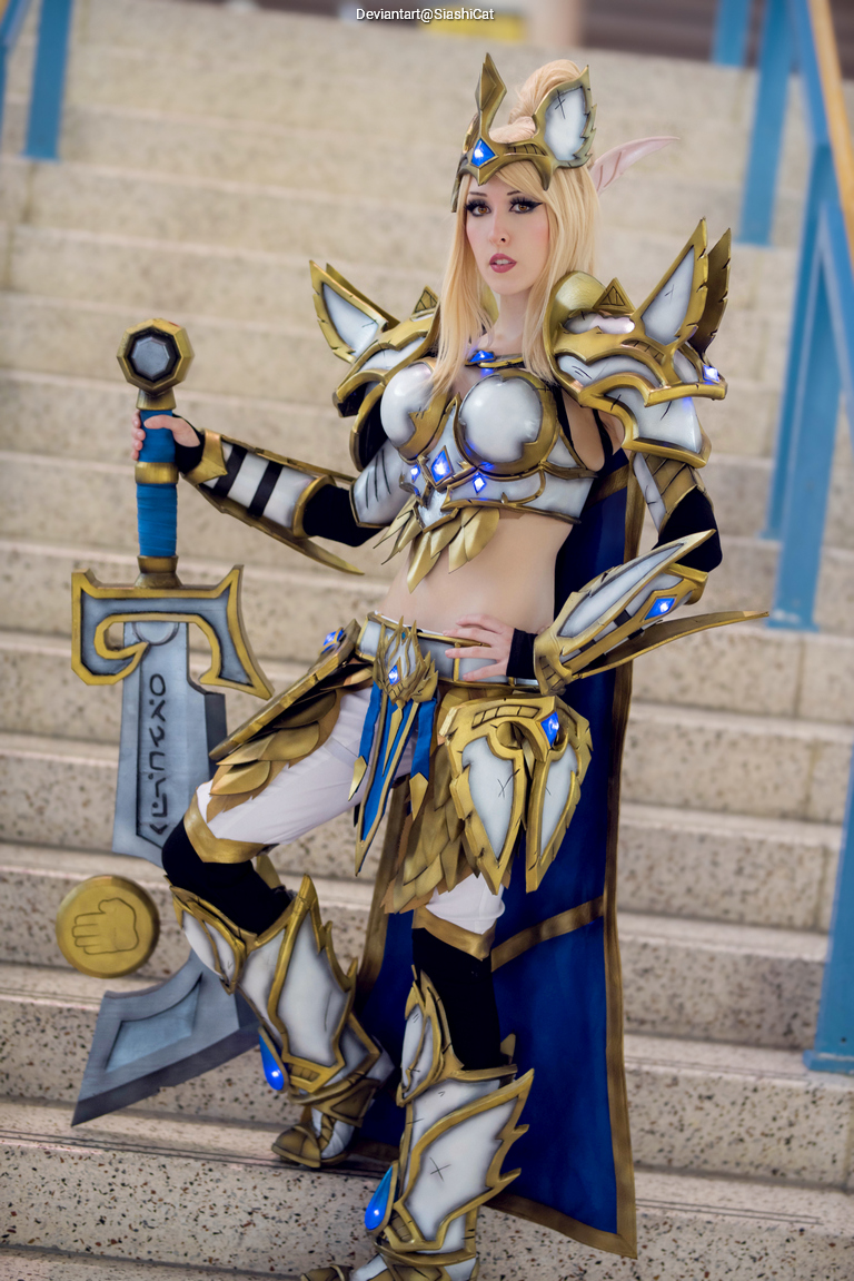 A person in elaborate fantasy armor stands confidently on a staircase, holding a sword decorated with blue gems and intricate designs.