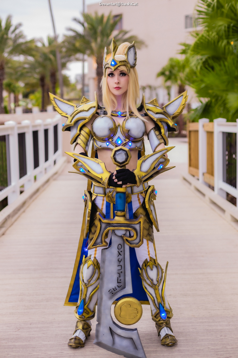 A person dressed in elaborate armor and holding a large sword, posing on a pathway lined with palm trees.