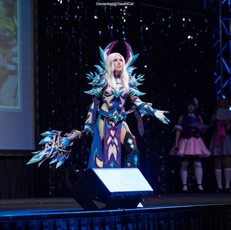 A person dressed in a detailed fantasy character costume with blue and purple armor, holding a large staff, posing on stage at an event.