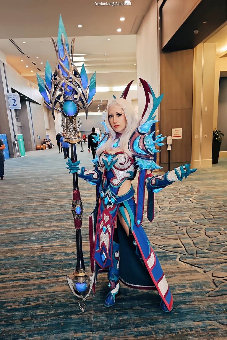 A cosplayer dressed as a character with long white hair, wearing a detailed armor set in blue and red tones, holding a large staff with intricate designs.