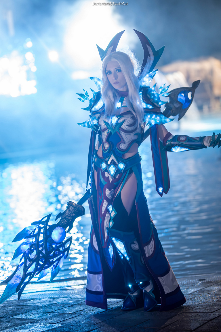 A person in an elaborate, blue and silver fantasy costume with glowing accents, standing by water with a magical staff in hand.