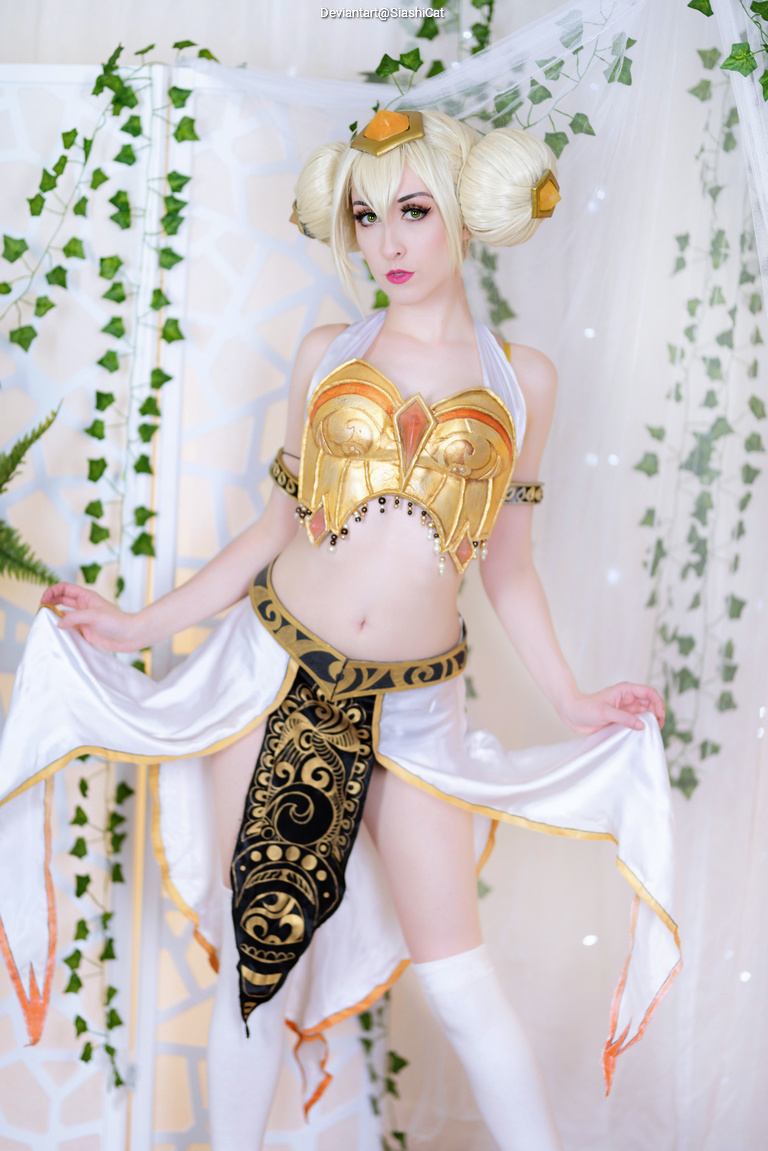 A person in a detailed fantasy costume featuring a gold and white top with ornate designs, paired with a flowing skirt and thigh-high stockings, posing amidst a decorative backdrop of draped fabric and green vines.