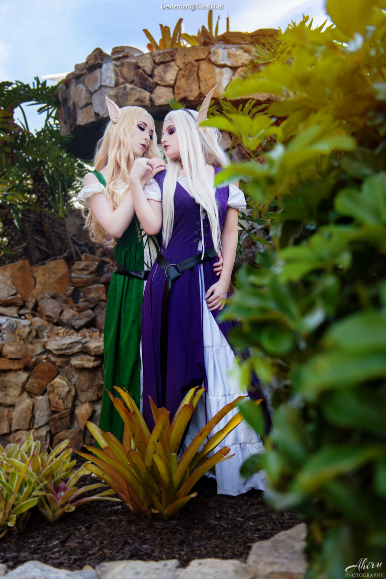 Two individuals dressed in colorful fantasy-themed costumes, standing close to each other amidst lush greenery and stone decorations.