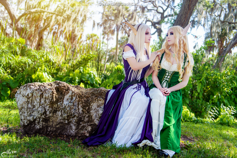 Two individuals in fantasy costumes sitting on a rock in a lush outdoor setting, one wearing a purple dress and the other in green, with elaborate hairstyles and makeup.
