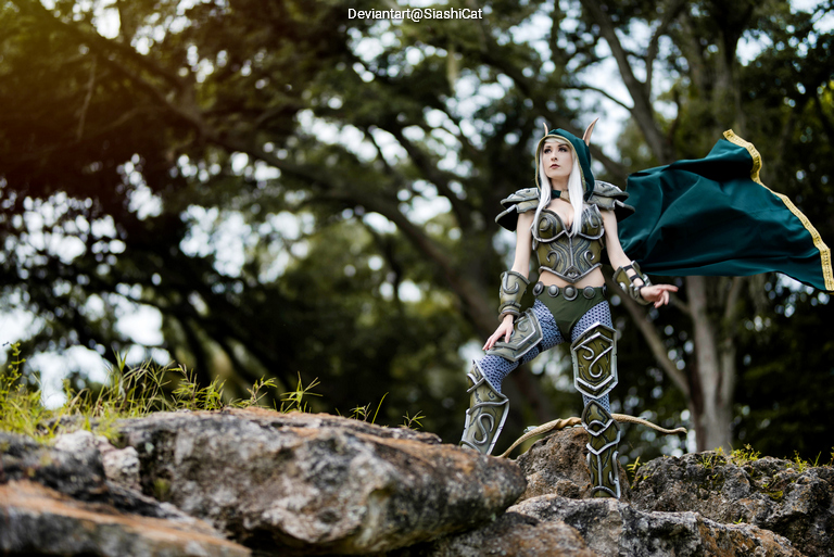 A cosplayer dressed in elaborate fantasy armor stands on a rock in a forested area, gazing into the distance.
