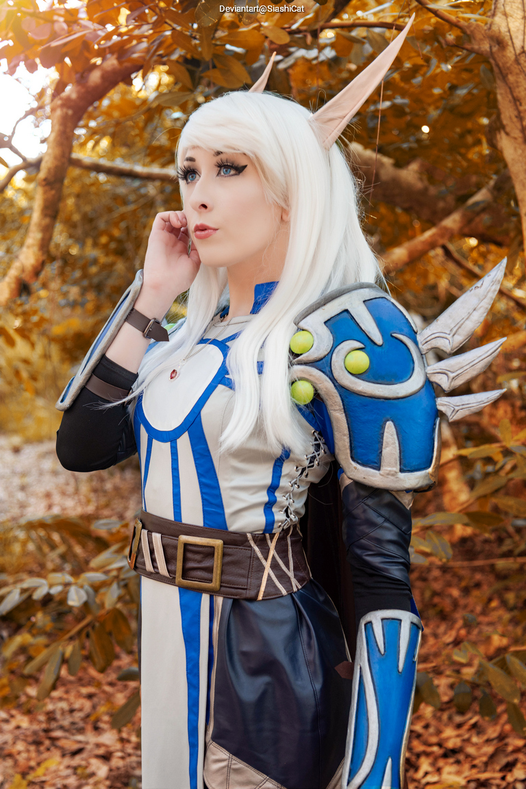 A person in a fantasy costume with white hair and pointed ears, standing in a wooded area with autumn leaves. The costume features a blue and silver design, emphasizing armor-like elements.