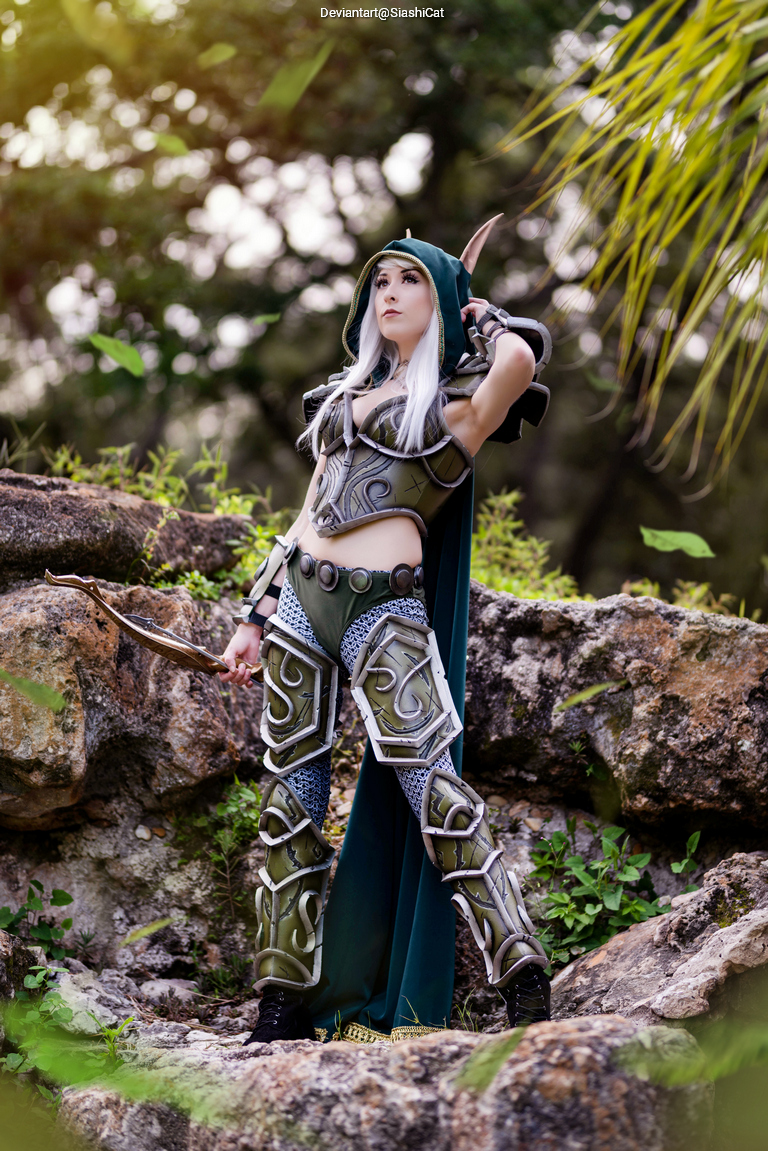 A female cosplayer dressed in an elaborate fantasy costume with armor and a cape, holding a bow and posing confidently on a rock amidst greenery.