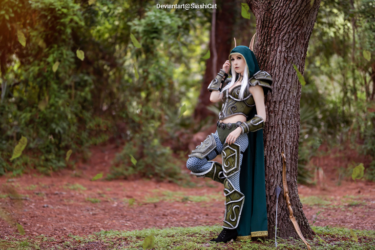 A person in an elaborate fantasy costume with armor and a green hood, standing next to a tree in a forested area.