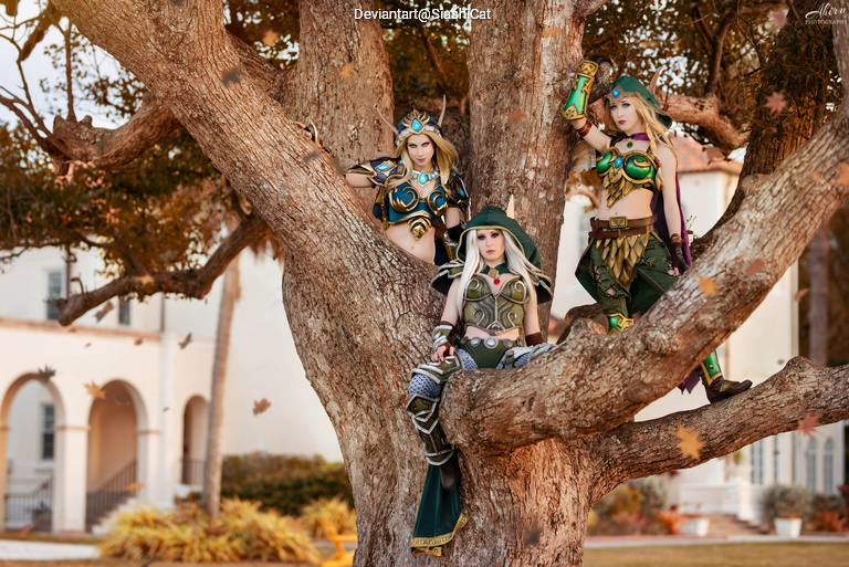 Three cosplayers dressed as fantasy characters pose in a tree, wearing elaborate costumes with armor and accessories. The setting includes a subtle garden background.