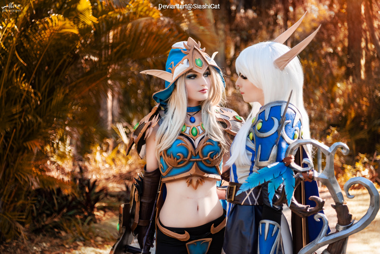 Two women in elaborate fantasy costumes engaged in conversation in a natural setting with greenery in the background.