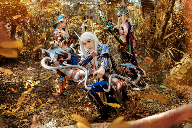 Three cosplayers in a forest, dressed in elaborate fantasy costumes, each holding a bow and arrows. The scene features autumn foliage, creating a vibrant backdrop.