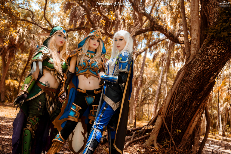 Three cosplayers dressed in elaborate fantasy costumes stand together in a wooded area, with sunlight filtering through the trees.