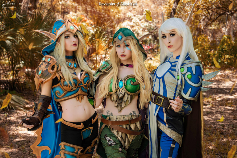 Three individuals in elaborate fantasy costumes, featuring intricate armor and accessories, posed together in a natural outdoor setting with colorful foliage in the background.
