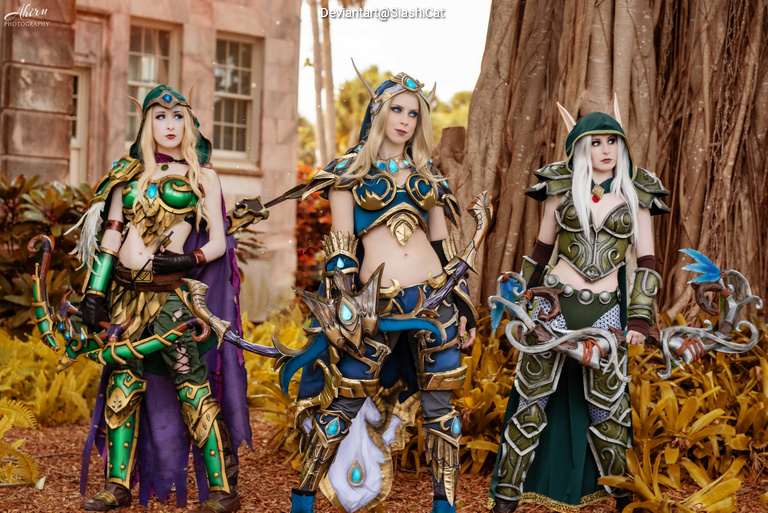 Three female cosplayers dressed as fantasy characters, standing in a lush outdoor setting. Each is wearing a detailed costume with intricate designs, showcasing colorful armor and accessories. The background features trees and plants, enhancing the vibrant atmosphere.