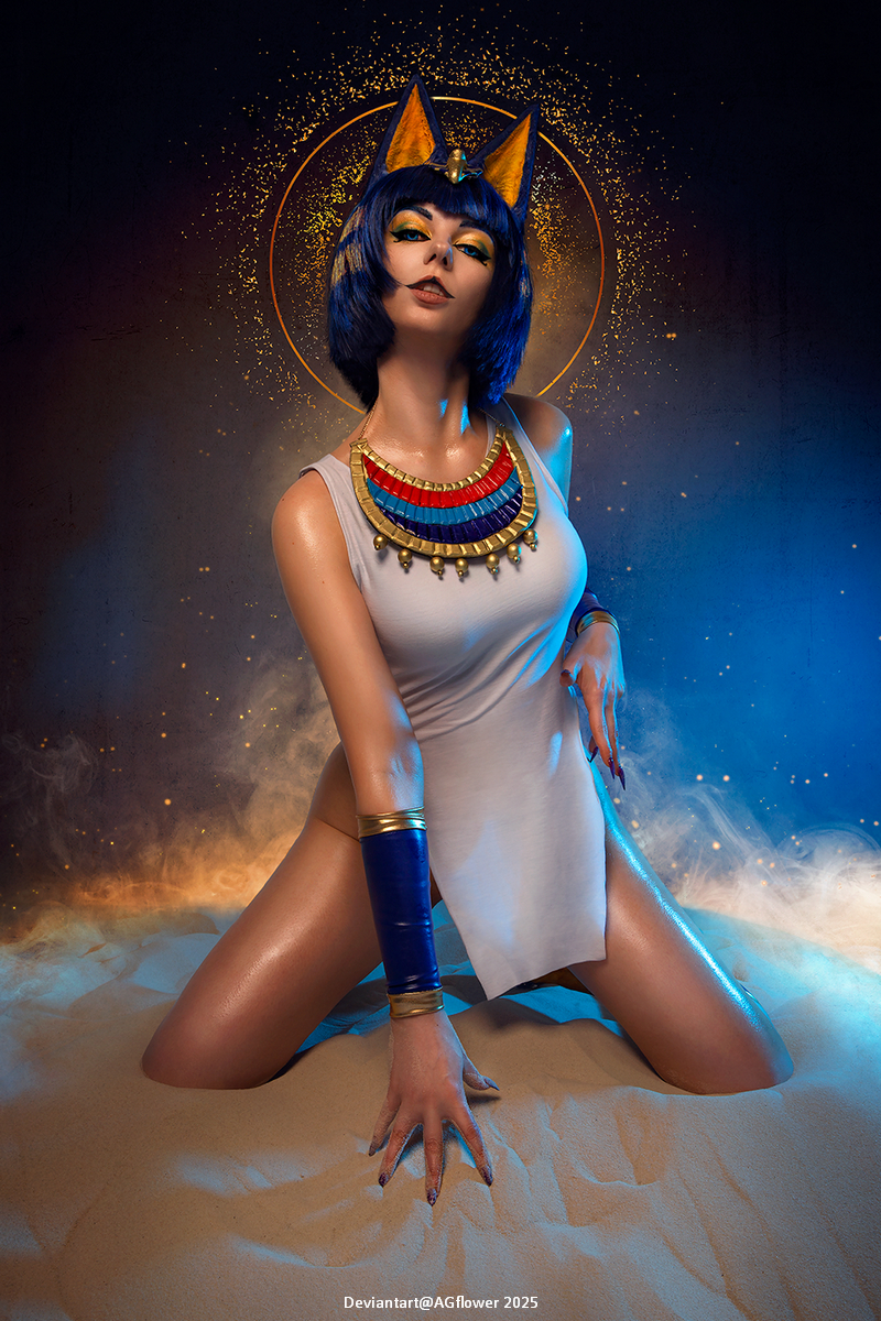 A person dressed as an Egyptian goddess, featuring a blue wig and ear-like headpieces, posed on sandy ground with golden accents in the background.