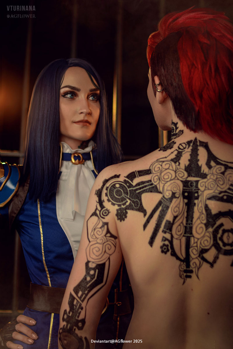 A close-up of two individuals, one in a blue outfit with long dark hair and the other with short red hair and intricate back tattoos. They are in a moment of connection, surrounded by dramatic lighting.