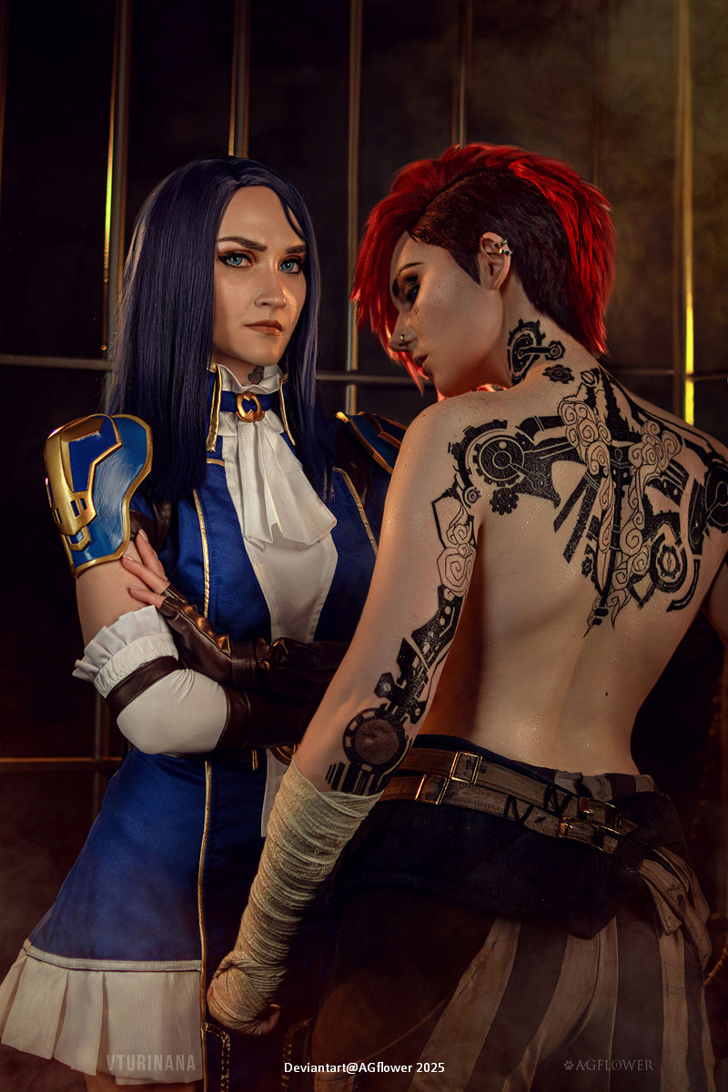 Two women in fantasy-themed costumes pose together. One has long dark hair and wears a blue and white outfit with gold accents, while the other has short red hair and tattoos on her back. The setting has dramatic lighting and a muted background.