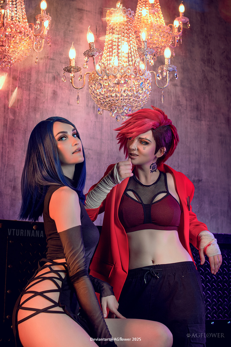 Two cosplayers posing under a sparkling chandelier. One has long blue hair and is wearing a black outfit, while the other has short red hair, a red jacket, and a sporty top.