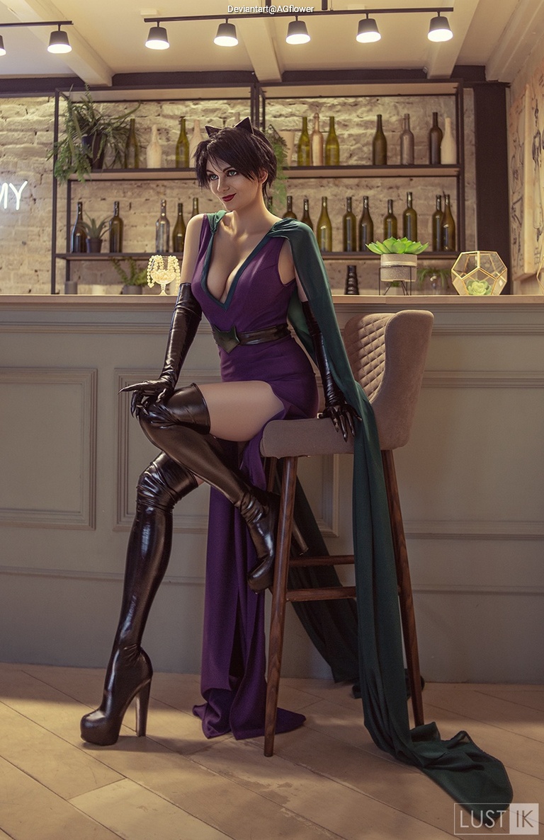 A woman in a stylish outfit with a purple dress and green cape, sitting on a bar stool in a modern interior with a shelf filled with bottles in the background.
