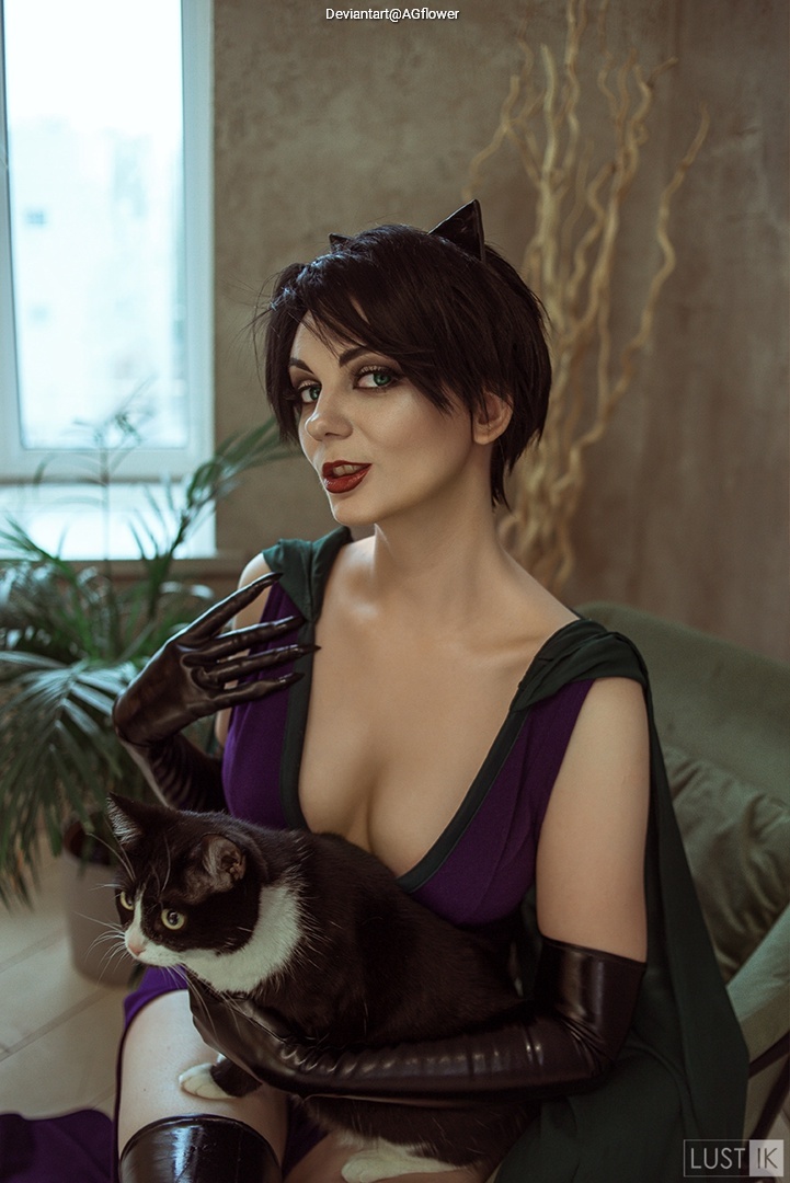 A woman dressed in a purple outfit with long black gloves, sitting on a green chair, holding a black and white cat in her lap. She has short dark hair with cat-like ears and is posing confidently in front of a window.