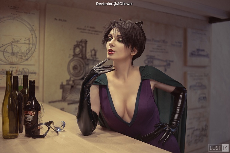 A woman in a costume with cat-like features sits elegantly at a bar, surrounded by several bottles. She has short, dark hair and wears a form-fitting purple outfit with a green cape, embodying a sultry character.