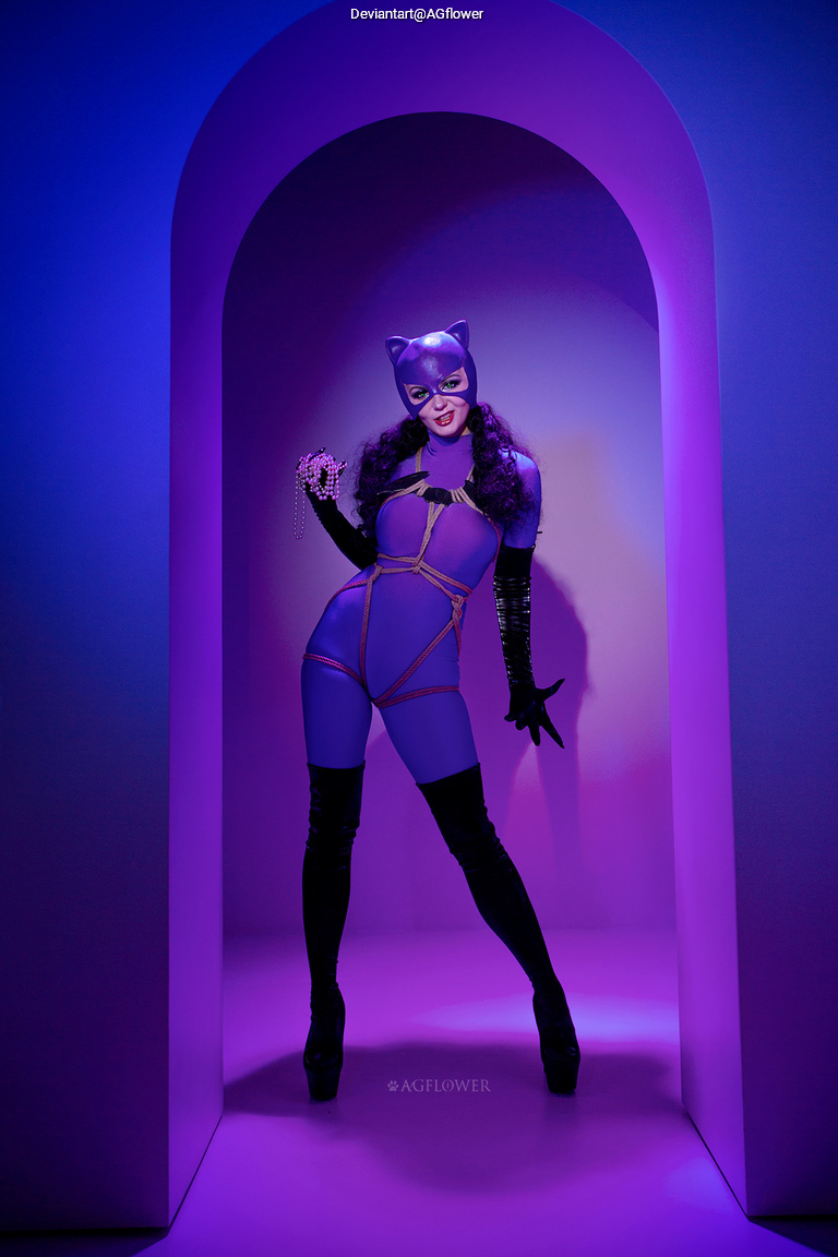 A person dressed in a purple catsuit and mask stands in a brightly lit archway, holding a string of pearls and posing confidently.