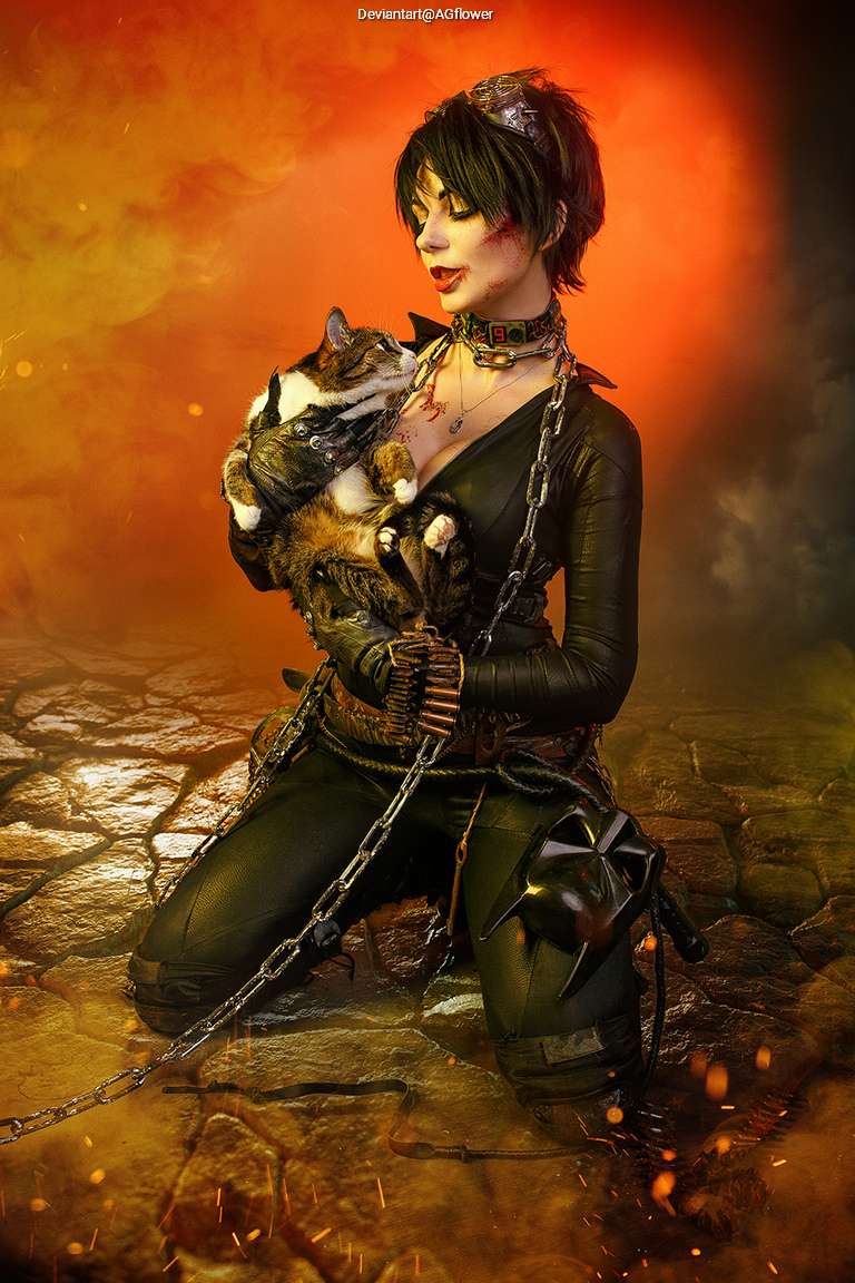 A woman in a dark outfit kneels on a rocky surface, holding a cat close while surrounded by a smoky, orange atmosphere.