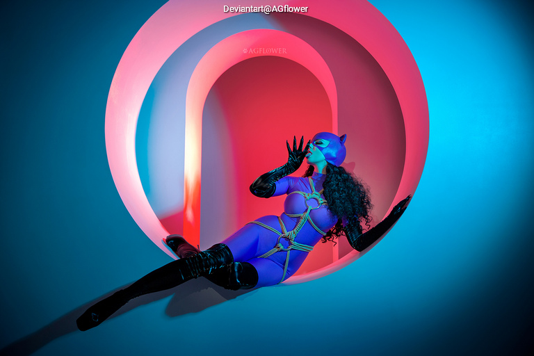 A person in a purple cat-like costume is posing gracefully in a circular cutout with vibrant blue and pink lighting in the background.